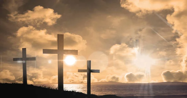 Image of three christian crosses over landscape with sun glowing on orange sky. easter celebration, religion and tradition concept digitally generated image.