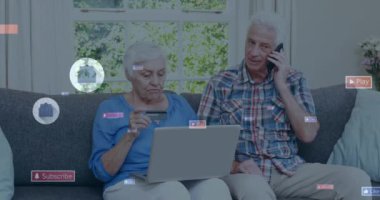 Animation of social media icons over caucasian senior couple using electronic devices at home. Social media networking technology concept