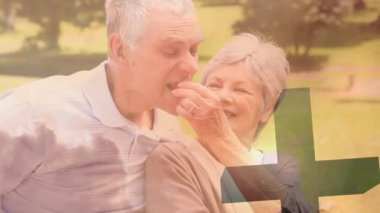 Animation of caucasian senior woman feeding strawberry to husband in park over silhouette cross. Catholicism, religion. Digital composite, multiple exposure, happy, love, together, picnic, retired.