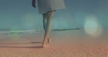 Animation of lens flare over low section of biracial young woman walking barefoot on sandy beach. Digital composite, vacations, leg, summer, nature and enjoyment concept.