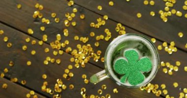 Video of st patrick's green shamrock in beer glass and gold sequins on wooden background. St patrick's day, irish tradition and celebration concept.