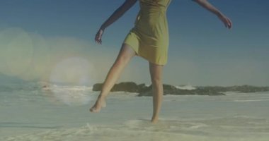 Animation of lens flares over biracial young woman with arms outstretched walking at shore. Digital composite, carefree, cheerful, vacations, sea, summer, nature and enjoyment concept.