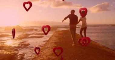 Animation of red hearts over couple in love holding hands by sea. Valentine's day, love and romance concept digitally generated video.