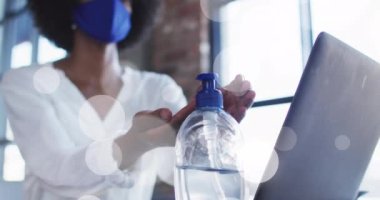 Animation of light spots over african american woman wearing face mask and disinfecting hands. Protection, virus, healthcare and lifestyle concept digitally generated video.
