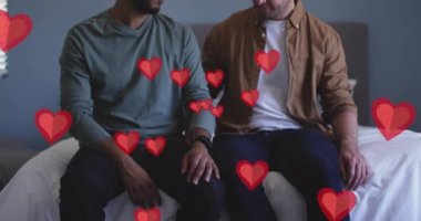Animation of red hearts over diverse male couple sitting on bed. Valentine's day, love and celebration concept digitally generated video.