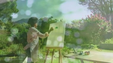 Animation of light spots over african american woman painting on easel in garden. Art, hobby and nature concept digitally generated video.
