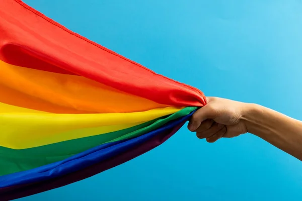 Hand holding rainbow coloured flag with copy space on blue background. Pride month, equality, lgbt and human rights concept.