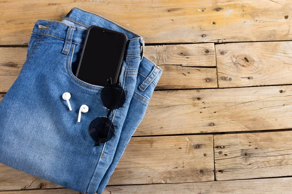 Blue jeans with smartphone, earphones and glasses lying on wooden surface. Clothes, fashion, design, fabrics, materials and shopping concept.