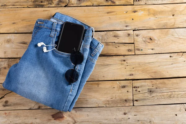 Blue jeans with smartphone, earphones and glasses lying on wooden surface. Clothes, fashion, design, fabrics, materials and shopping concept.