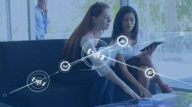 Animation of connected icons over diverse female colleagues discussing reports in office. Digital composite, multiple exposure, business, communication, teamwork and technology concept.