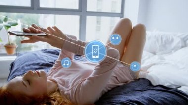 Evdeki yatakta uzanan akıllı telefonu kullanan beyaz kadın üzerindeki dijital simge ağının animasyonu. Küresel ağ ve iş teknolojisi kavramı