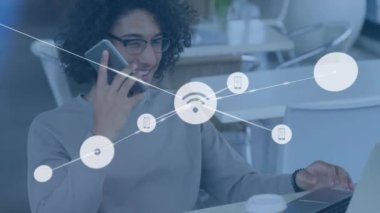 Animation of connected icons over biracial man talking over phone and working on laptop in cafes. Digital composite, multiple exposure, communication, wifi, phone, business and technology concept.