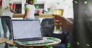 Animation of clover icons over diverse friends drinking beer and using laptop with copy space. St patricks day and celebration concept digitally generated video.