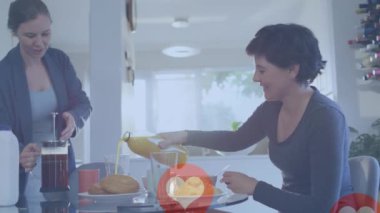 Animation of heart emojis and numbers over happy caucasian female couple in love at home. Love, romance and celebration concept digitally generated video.