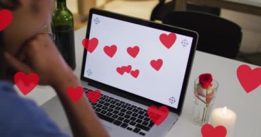 Animation of hearts over biracial man drinking wine and using laptop with copy space. Valentines day and celebration concept digitally generated video.