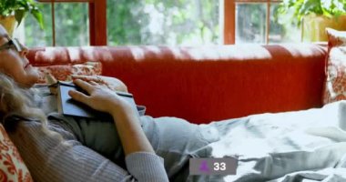Animation of social media icons over caucasian senior woman holding a book sleeping on the couch. Social media networking technology concept