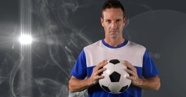 Caucasian male soccer player holding football against smoke effect and light spot on grey background. sports tournament and competition concept