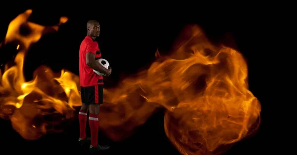 Composition of male football player standing holding ball over flames on black background. sport and competition concept digitally generated image.