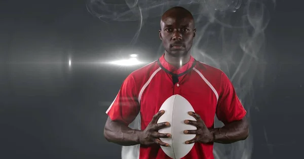 African american male rugby player holding ball against smoke and light spot on grey background. sports tournament and competition concept