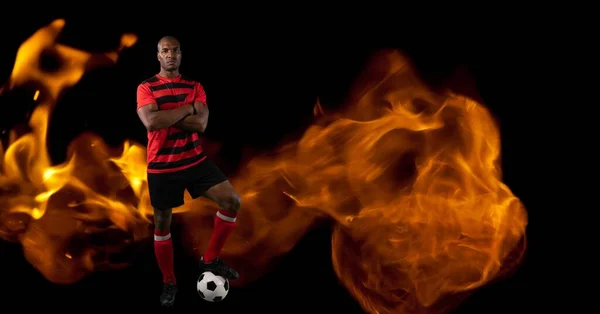 Composition of male football player standing with ball over flames on black background. sport and competition concept digitally generated image.