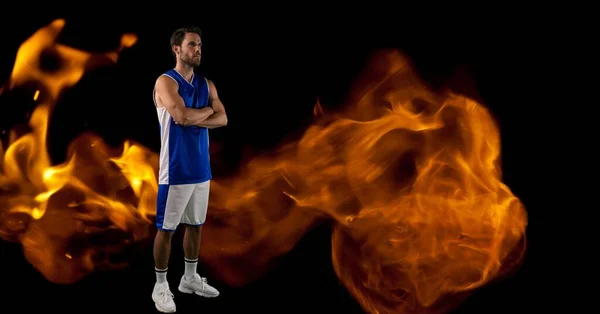 Composition of male basketball player standing with arms crossed over flames on black background. sport and competition concept digitally generated image.