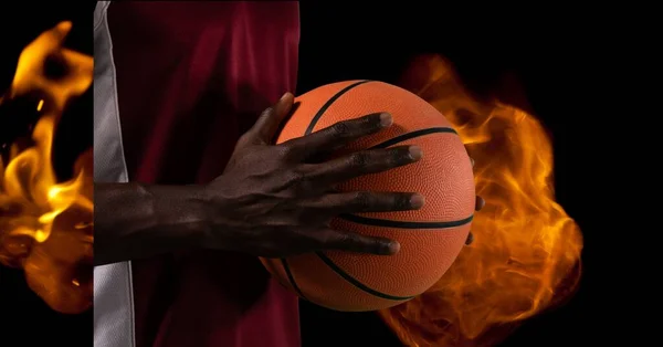 Composition of midsection of male basketball player holding ball over flames on black background. sport and competition concept digitally generated image.