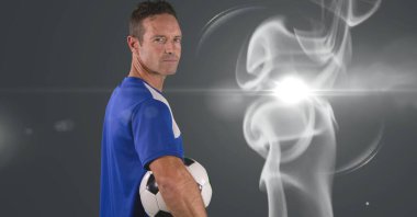 Caucasian male soccer player holding football against smoke effect and light spot on grey background. sports tournament and competition concept