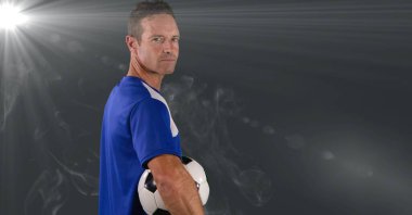 Caucasian male soccer player holding football against smoke effect and light spot on grey background. sports tournament and competition concept
