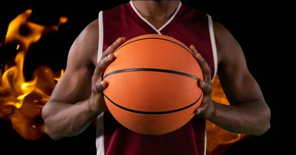 Composition of midsection of male basketball player holding ball over flames on black background. sport and competition concept digitally generated image.