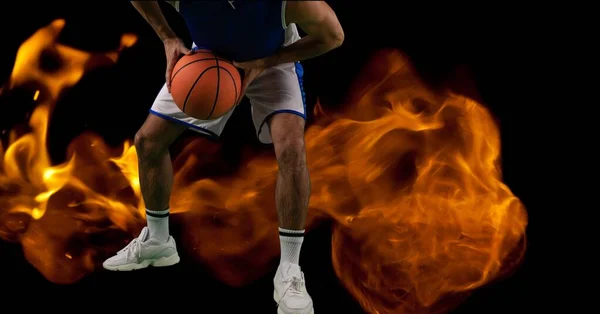 Composition of low section of male basketball player holding ball over flames on black background. sport and competition concept digitally generated image.