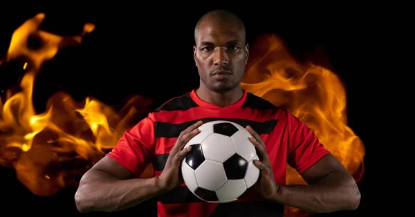 Composition of male football player holding ball over flames on black background. sport and competition concept digitally generated image.