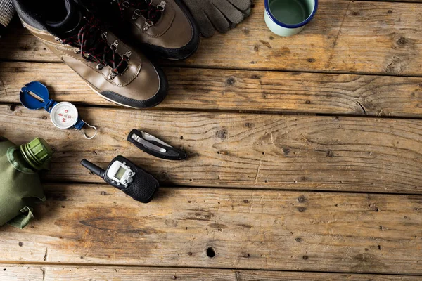 Camping equipment of trekking shoes, metal cup and compass on wooden background with copy space. National camping month, equipment and celebration concept.