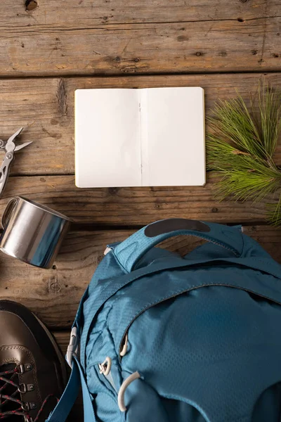Camping equipment of backpack, metal cup and blank notebook on wooden background with copy space. National camping month, equipment and celebration concept.