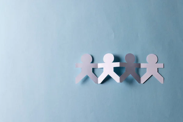 Close up of four paper cut out people figures holding hands with copy space on blue background. Humanitarian, people, help and human concept.