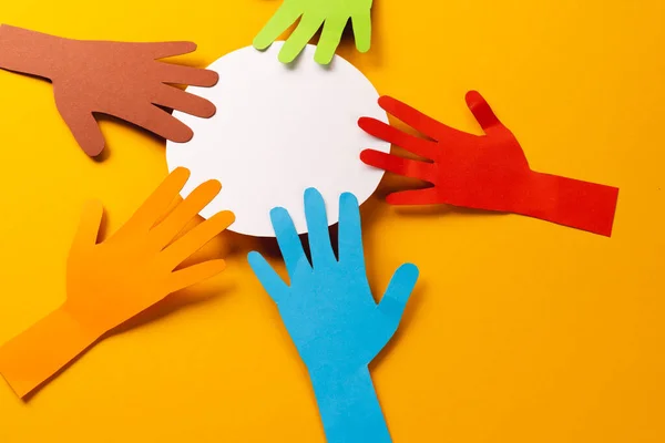 Paper cut out of multi coloured hands and white circle with copy space on yellow background. Humanitarian, people, help and human concept.