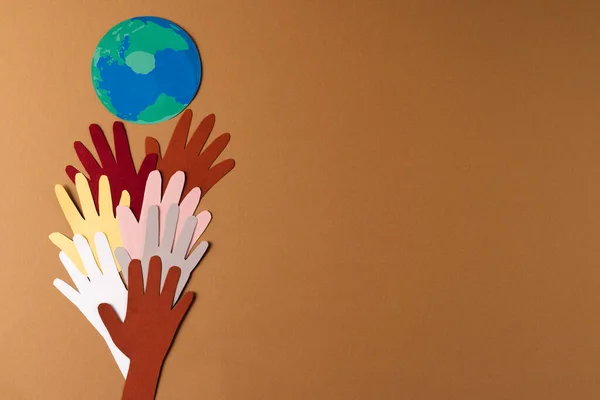 Paper cut out of multi coloured hands and globe with copy space on brown background. Humanitarian aid, people, help and human concept.