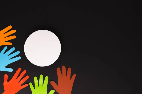 Paper cut out of multi coloured hands and white circle with copy space on black background. Humanitarian aid, people, help and human concept.