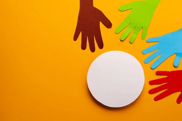 Paper cut out of multi coloured hands and white circle with copy space on orange background. Humanitarian aid, people, help and human concept.