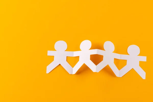 Close up of four paper cut out people figures holding hands with copy space on yellow background. Humanitarian, people, help and human concept.