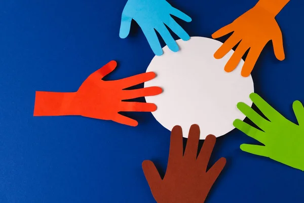 Paper cut out of multi coloured hands and white circle with copy space on blue background. Humanitarian, people, help and human concept.
