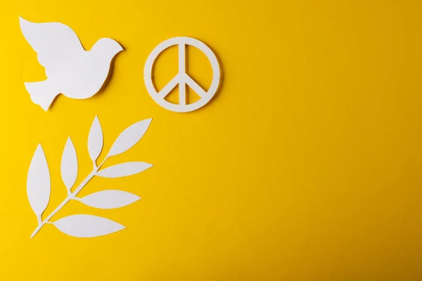 High angle view of white peace sign, white dove and leaf with copy space on yellow background. Peace and anti war movement concept.