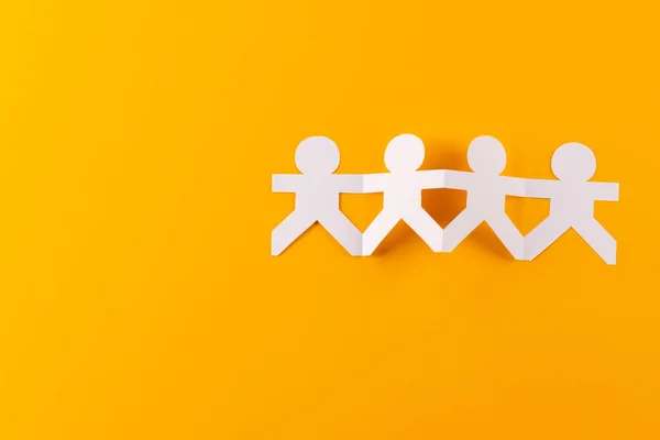 Close up of four paper cut out people figures holding hands with copy space on orange background. Humanitarian, people, help and human concept.
