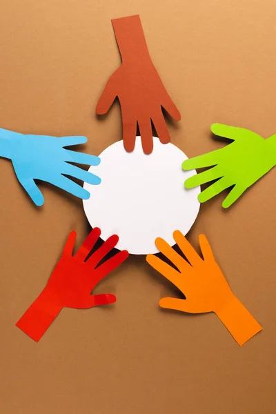 Paper cut out of multi coloured hands and white circle with copy space on brown background. Humanitarian, people, help and human concept.