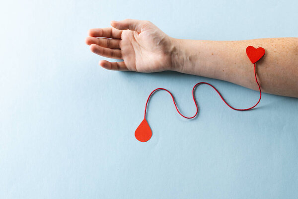 Arm of caucasian woman with heart and red string with blood drop, on blue background. Blood donation, medicine and healthcare.