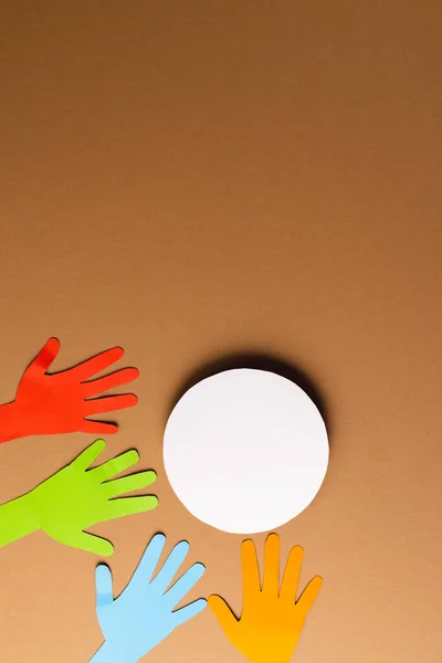 Paper cut out of multi coloured hands and white circle with copy space on brown background. Humanitarian aid, people, help and human concept.