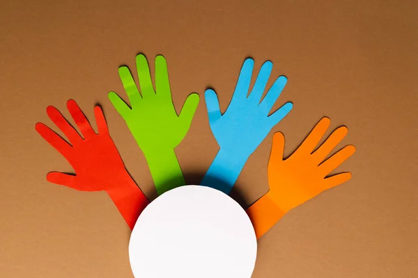 Paper cut out of multi coloured hands and white circle with copy space on brown background. Humanitarian aid, people, help and human concept.