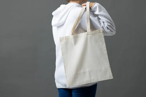 Caucasian woman holding over the shoulder white canvas bag with copy space on grey background. Bags and fashion concept.