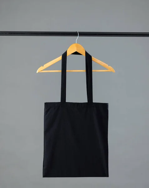 Close up of black canvas bag hanging on wooden clothes hanger with copy space on grey background. Eco friendly shopping bags and fashion concept.