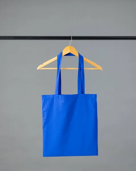 Close up of blue canvas bag hanging on wooden clothes hanger with copy space on grey background. Eco friendly shopping bags and fashion concept.
