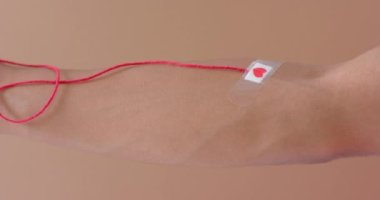 Kan damlası olan melez bir adam, kırmızı ip ve kalp bej üzerine alçı yapıştırıyor, ağır çekimde. Kan bağışı, ilaç ve sağlık hizmetleri.